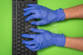 Closeup of black computer keyboard and hands in blue rubber gloves. Work from home covid-19 concept.