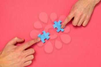 Hands of woman and man matching pieces of blue jigsaw puzzle on pink background. Relationship concept