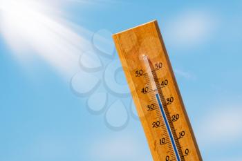 Thermometer with a high temperature reading on a scale, against a background of bright sun and a blue sky with clouds. The concept of hot, dangerous weather, global warming.