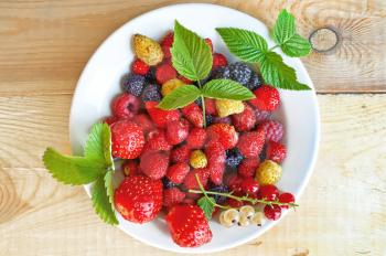 Strawberry, raspberry, blackberry, white and red currant, green leaves of strawberries and raspberry in white bowl on wooden boards