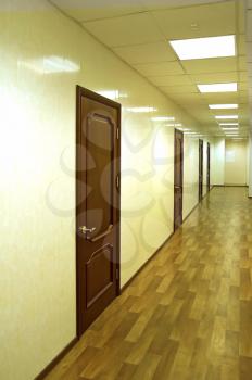 Several brown door in the hallway at the office against the beige wall, lamps on the ceiling
