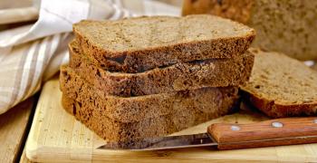 A stack of slices of rye homemade bread on a board with a knife, napkin, loaf of bread on a wooden board