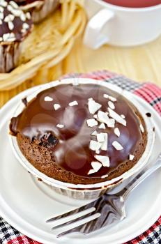 Chocolate cake, fork on the plate and red napkin, cup of tea, wicker plate of cupcakes on the background of wooden boards