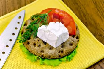 A piece of feta cheese, a slice of tomato, dill on a round rye crispbread with lettuce, a knife and a yellow napkin on the background of wooden board
