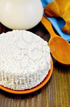 Cottage cheese in a saucer, wooden spoon, a bagel, a pitcher of milk, napkin on wooden board