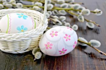 Two Easter eggs painted with a pattern in the form of flowers and stripes, white basket, willow twigs on the background of wooden boards