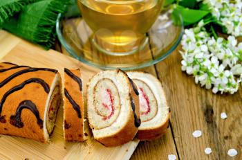 Biscuit roulade with cream and jam, a glass cup with tea, napkin, flowers and leaves of bird-cherry on a background of wooden boards