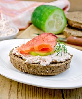 Sandwich of rye bread with cream, cucumber, dill and salmon on a white plate on a wooden board