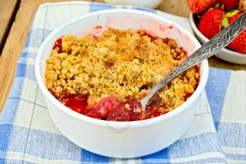 Strawberry crumble in a white bowl with a spoon on a napkin, strawberries on a wooden boards background