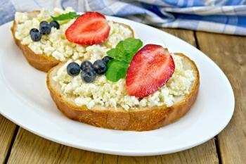 Slices of bread with curd cream, mint, blueberries and strawberries on a plate on a wooden boards background