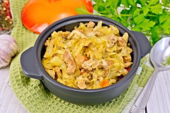 Cabbage stew with meat in a black pot on a napkin, cover, garlic, pepper, parsley. spoon on a background of light board