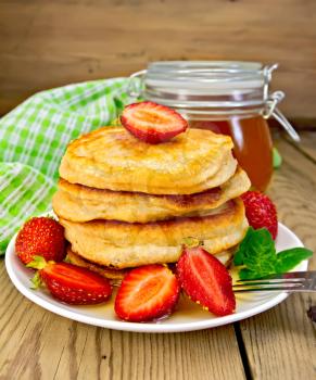 A stack of pancakes with strawberries and honey on a white plate, a jar of honey, a napkin on a wooden boards background