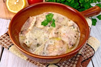 Fish stew with cream sauce in a ceramic pan, lemon, parsley, tomato, napkin on background light wooden boards