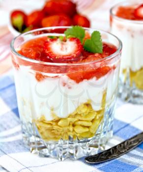 Dairy dessert with strawberries, corn flakes and yogurt in two glassful, spoon, blue napkin, strawberries in a bowl on linen tablecloth background