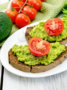 Two slices of rye bread with guakomole and tomato on a plate, napkin, avocado on a background of light wooden boards