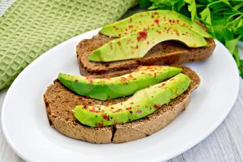 Two slices of rye bread with slices of avocado and pepper on a white plate, green napkin, parsley on the background light wooden boards