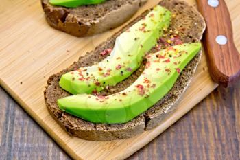 Two slices of rye bread with slices of avocado and pepper, knife against the dark wooden board