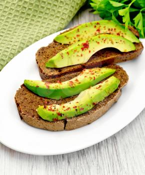 Two slices of rye bread with slices of avocado and pepper on a plate, napkin, parsley on the background light wooden boards