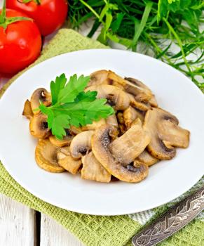 Champignons fried in a plate with parsley leaf, fork, tomato on a napkin on the background light wooden boards