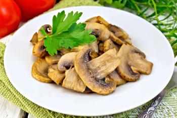 Champignons fried in a plate with parsley leaf, fork, ripe tomatoes on a napkin on the background light wooden boards