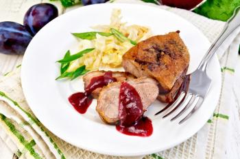 Roasted duck breast with braised cabbage, green onions and plum sauce in a white plate on a towel, plum and fork on the background light wooden boards