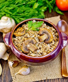 Boiled buckwheat with mushrooms in a brown pottery bowl on a napkin of burlap, parsley, tomatoes and garlic on a wooden boards background