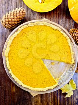 Pumpkin pie in a glass pan, pumpkin, cones and maple leaf on a sacking on a background of dark wooden boards on top