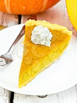 A piece of pumpkin pie with whipped cream, a fork in the white plate, pumpkin on the background light wooden boards