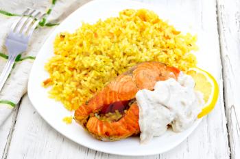 Pink salmon with lemon, milk sauce and rice with turmeric in a dish, a towel and a fork on the background light wooden boards