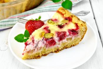 Piece of pie with strawberries, rhubarb and cream sauce, fork, berries and mint in white plate, a frying pan with pie on a napkin on a wooden boards background