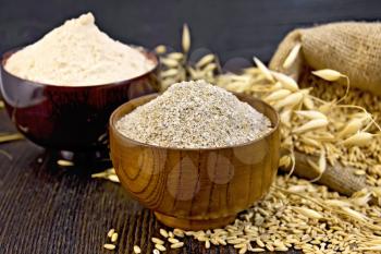 Bran flakes oat and oat flour in two wooden bowl, a bag of grain oats, oat ears against the dark wooden board