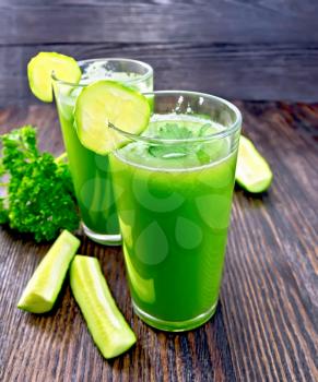 Cucumber juice in two tall glass with slices of cucumber, cucumber and parsley on a wooden board background