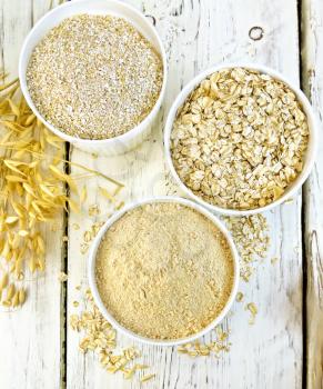 Flour oat, bran and oat flakes in three white bowls, stalks of oats on the background of the wooden planks on top