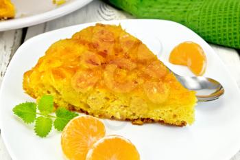 Piece of tart  with mandarin, mint, tangerine slices and spoon in white plate, a towel on the background light wooden boards