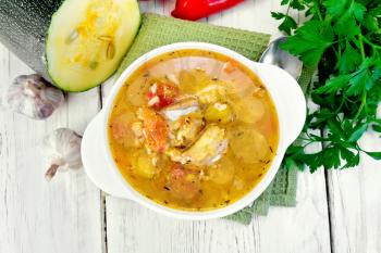 Fish soup with zucchini, peppers and rice in a white bowl on a napkin, garlic, parsley on a background of wooden boards on top