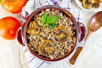 Boiled buckwheat with champignon in a brown pottery bowl on a kitchen towel, tomatoes in the background light from the top of the board