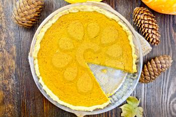 Pumpkin pie in a glass pan on sackcloth, cones and maple leaf on the background of the wooden planks on top