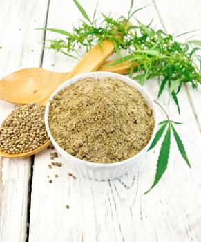Hemp flour in white bowl, grain in spoon, cannabis leaves on the background of the wooden boards