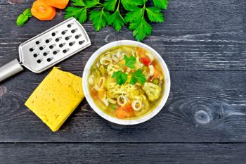 Minestrone soup with meat, celery, tomatoes, zucchini and cabbage, green peas, carrots and pasta in a white bowl, cheese, grater on background black wooden plank on top