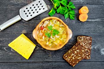 Minestrone soup with meat, celery, tomatoes, zucchini and cabbage, green peas, carrots and pasta in an earthenware bowl, cheese, grater on black background wooden plank on top