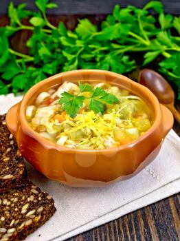 Minestrone soup with meat, celery, tomatoes, zucchini and cabbage, green peas, carrots and pasta in an earthenware bowl on a napkin, cheese and bread on a wooden boards background
