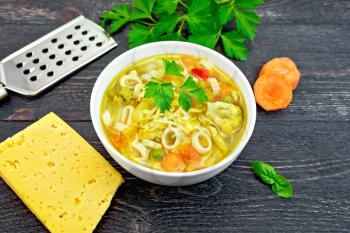 Minestrone soup with meat, celery, tomatoes, zucchini and cabbage, green peas, carrots and pasta in a white bowl, cheese, grater on background black wooden plank