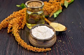 Flour amaranth in a clay bowl, oil in glass jar, a spoon with grain, brown flower with green leaves on a background of wooden boards