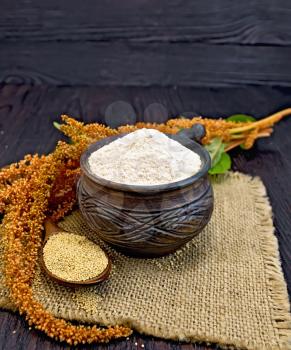 Flour amaranth in a clay cup, a spoon with grain, brown flower with green leaves on a napkin from a sacking on a background of dark wood planks