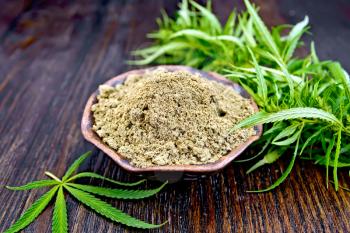 Hemp flour in a bowl, green cannabis leaves on the background of dark wooden boards