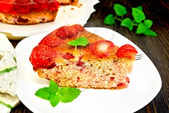 Piece of sweet pie with strawberries, kissel, jelly and mint, fork in white plate, napkin on a dark wooden board