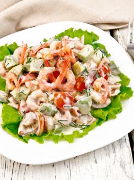 Salad with shrimp, avocado, tomatoes and mayonnaise on the green lettuce in a white plate, napkin, fork on the background light wooden boards