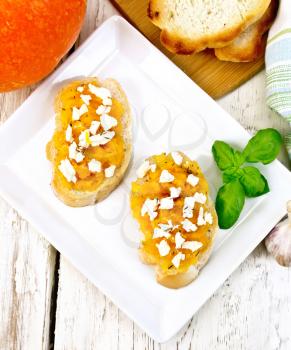 Bruschetta with pumpkin, salted feta cheese in a plate, garlic, basil and parsley, napkin and vegetable on a wooden board background from above