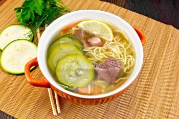 Soup with zucchini, beef, ham, lemon and noodles in a bowl, parsley and dill on a bamboo napkin against a dark wooden board