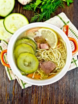 Soup with zucchini, beef, ham, lemon and noodles in a red bowl, parsley and dill on a napkin on a wooden board background on top